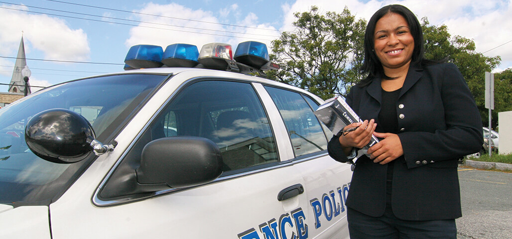 A Law Enforcement Certificate student stands, with books in hand, outside next to a police car.