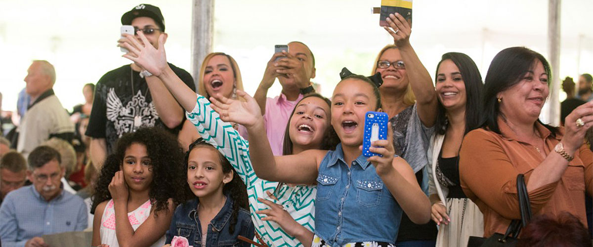 A group of people at commencement including children, standing up, cheering and waving.