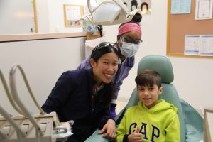 Dental patient Gustavo Rocha sits in a chair, giving the thumbs-up sign, while Dr. Bridget Ko and NECC student Rosa Livingston stand off to the left.