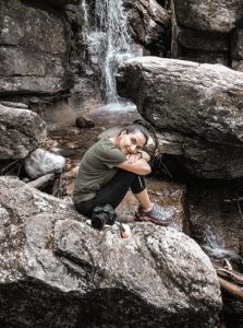 Alejandra sits on a rock in front of a waterfall with her camera at her side. 