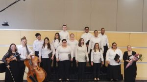 NECC Chorus students stand with black pants and white tops holding the selections of music to perform