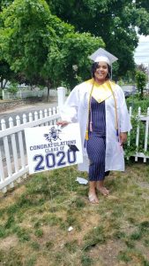 2020 grad stands in front of picket fence wearing her cap and gown