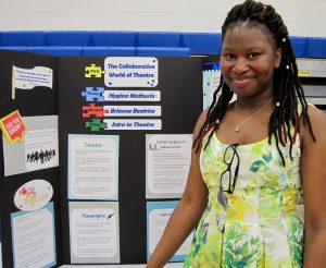 Young Black woman in floral dress stands in front of her honors project titled The Collaborative World of Theater.