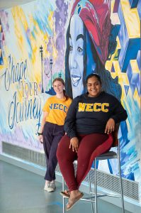 Two young woman wearing NECC t-shirts in front of a colorful mural on the Lawrence Campus.