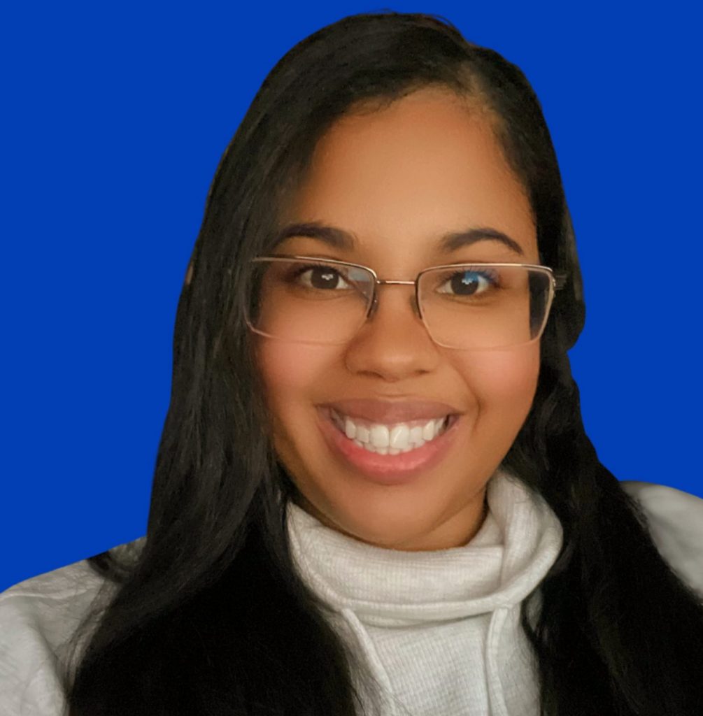 Ana, wearing glasses and a turtleneck sweater, smiles at camera in front of blue backdrop