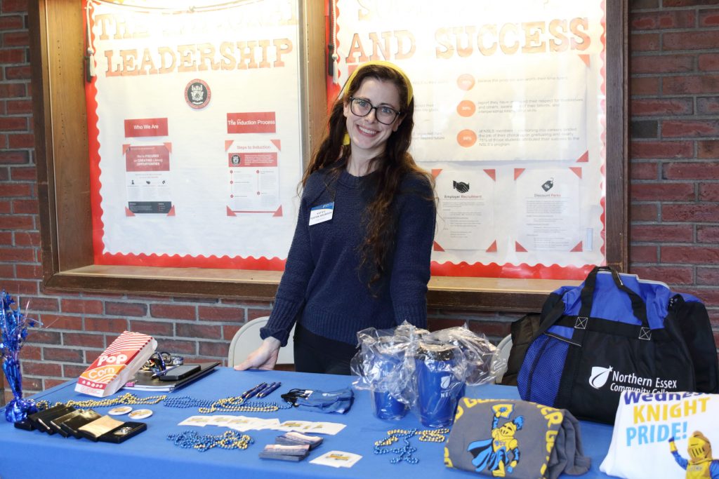 Katie stands behind table covered in blue table cloth, with various NECC swag items displayed