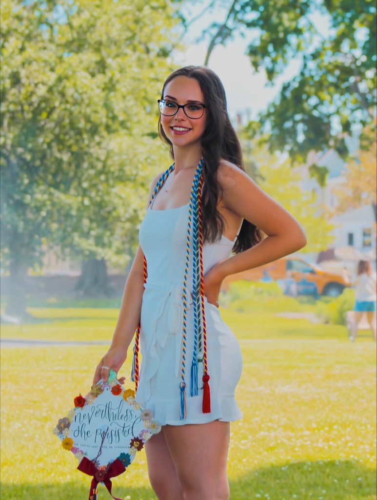Sierra stands smiling with her hand on her hip and graduation cap in other hand