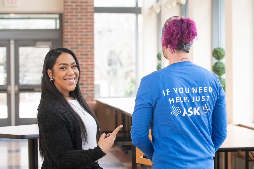 Chucha poses with fellow student ambassador, points to shirt that says "if you need help, just ask"