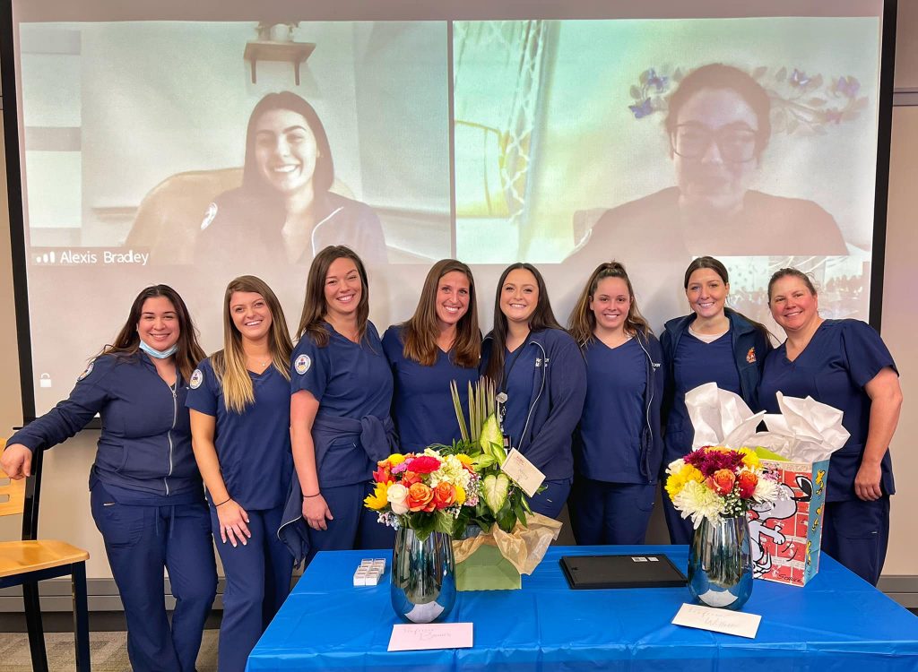 group of 10 students pose for picture in front of projection screen with images of two other students. All wear blue scrubs
