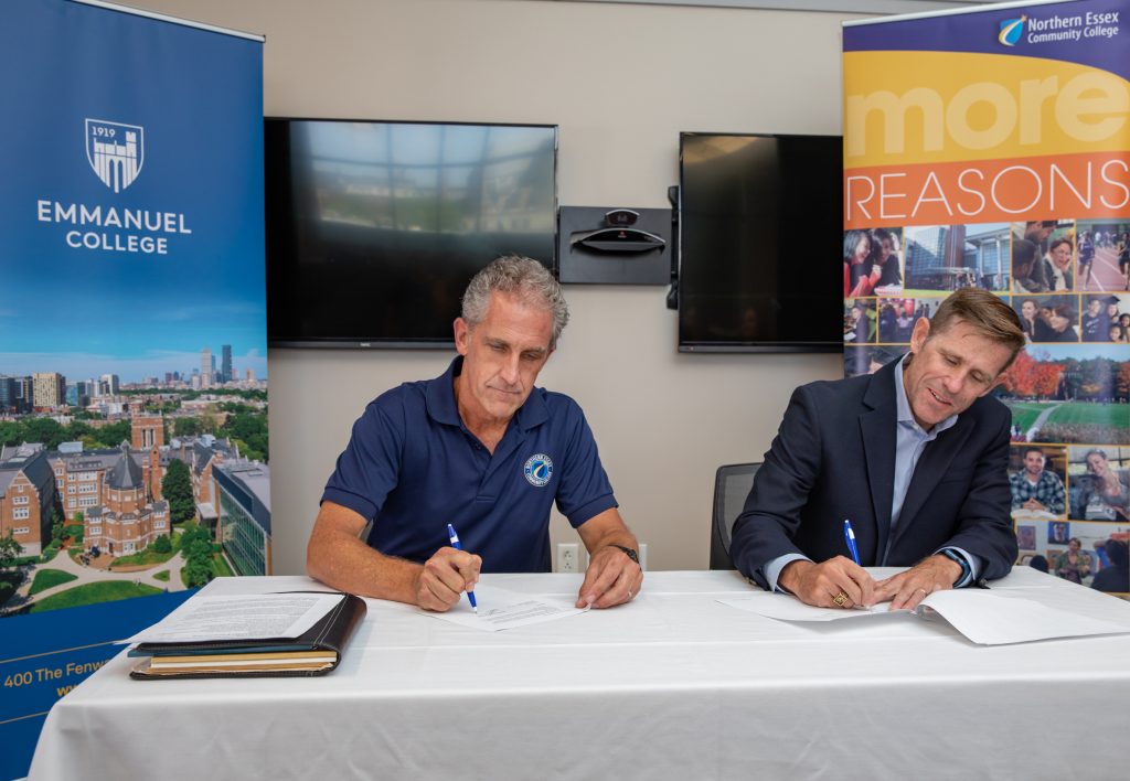 Dr Glenn and Emmanuel Rep sign documents in front of NECC and Emmanuel College Banners