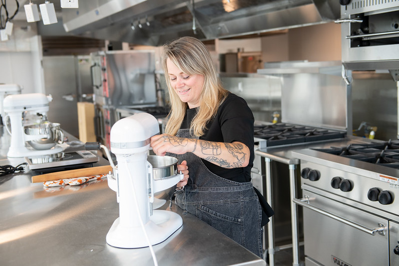 woman stands in commercial kitchen adding mixture to white kitchenaid mixer