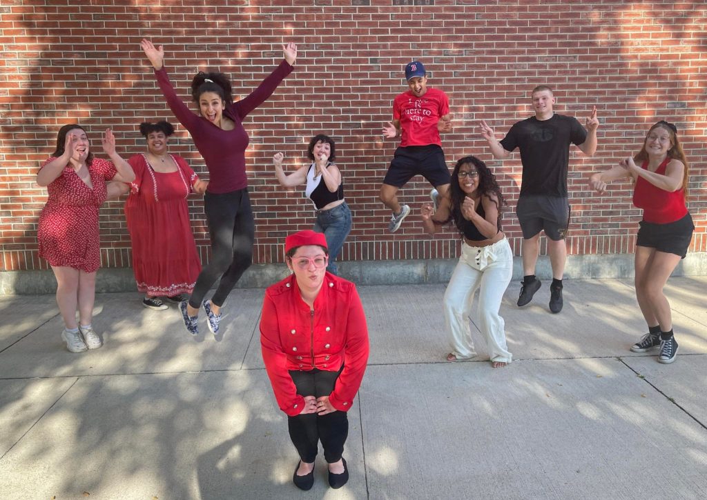 nine members of the cast jump up in unison in front of a brickwall