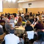 Wide shot of room full of tables and people seated, looking forward at large presentation screen