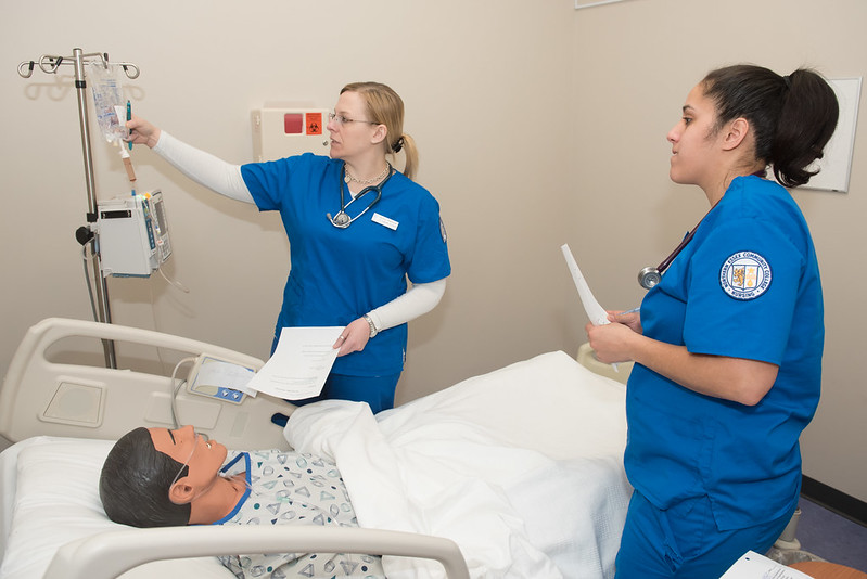 two nursing students look at IV bag next to hospital bed containing life-life training dummy