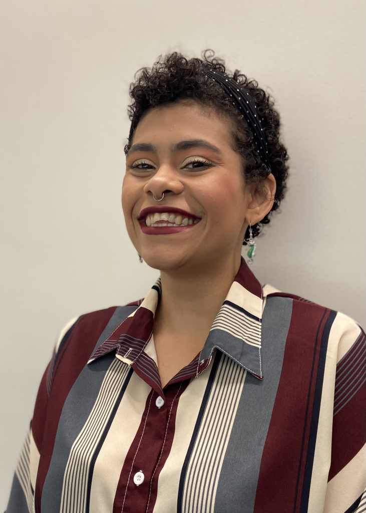 sarah stands smiling at the camera against an off-white wall