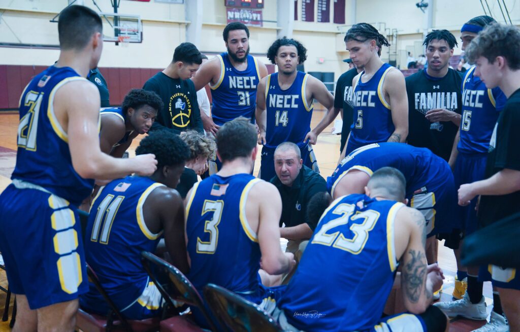 necc basketball team huddles up during a recent game. players sit and stand surrounding their coach