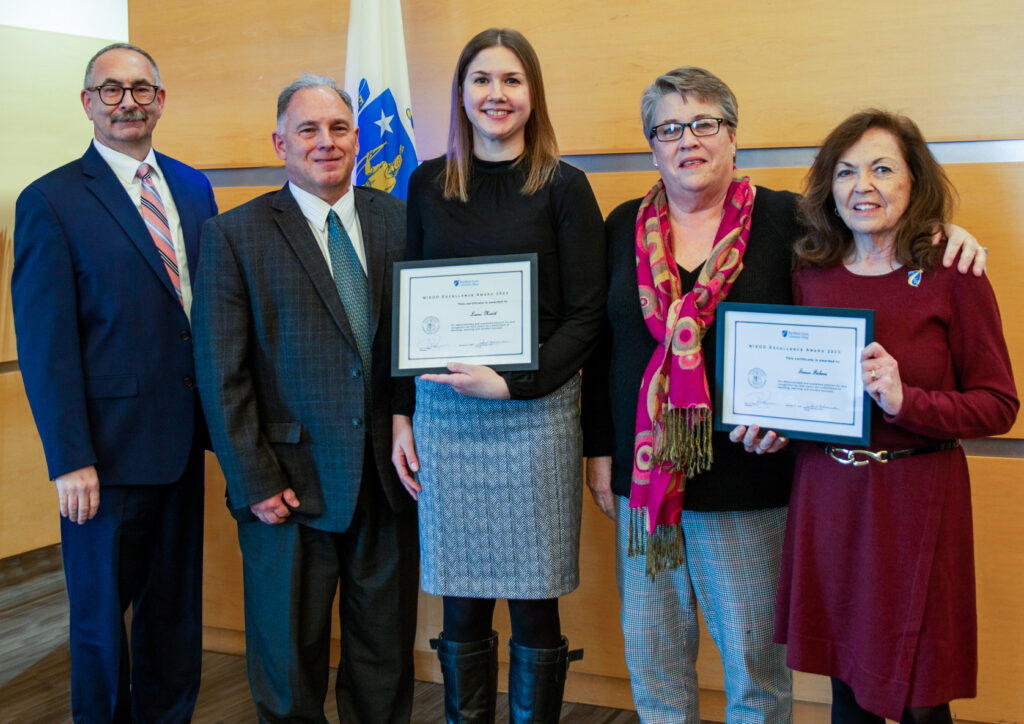 five people pose in front of wall, two hold paper awards