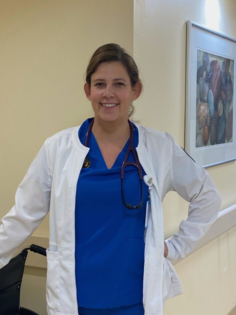 Karen poses in bright blue scrubs and white lab coat in hospital hallway