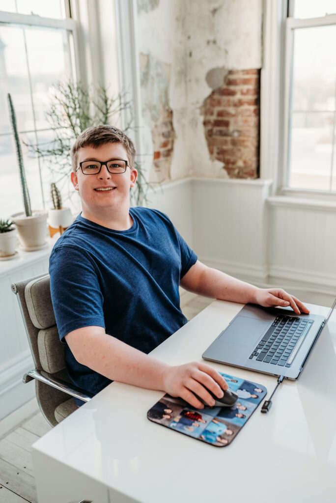 Shaun Hood sits at a desk with a laptop computer and smiles at the camera