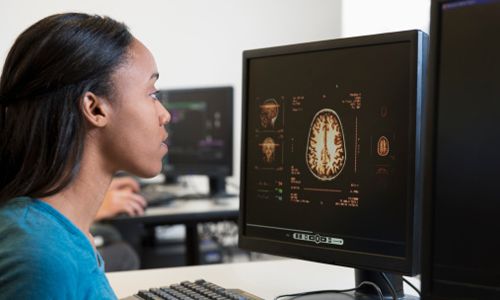 A woman sits at a computer looking at a complicated brain scan.