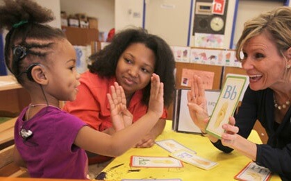 A little girl with a cochlear implant and two adults are practicing flashcards and signing B in ASL.