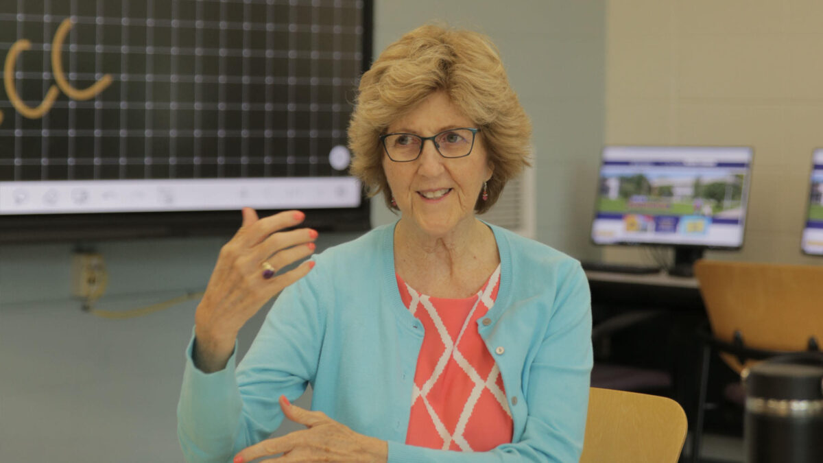 An image of a female presenting NECC tutor raising her arm while talking to someone out of frame in a classroom setting