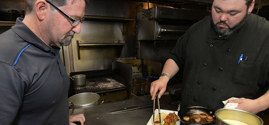 A student in the Hospitality Management associate degree program oversees a chef as he lays out food on a tray.