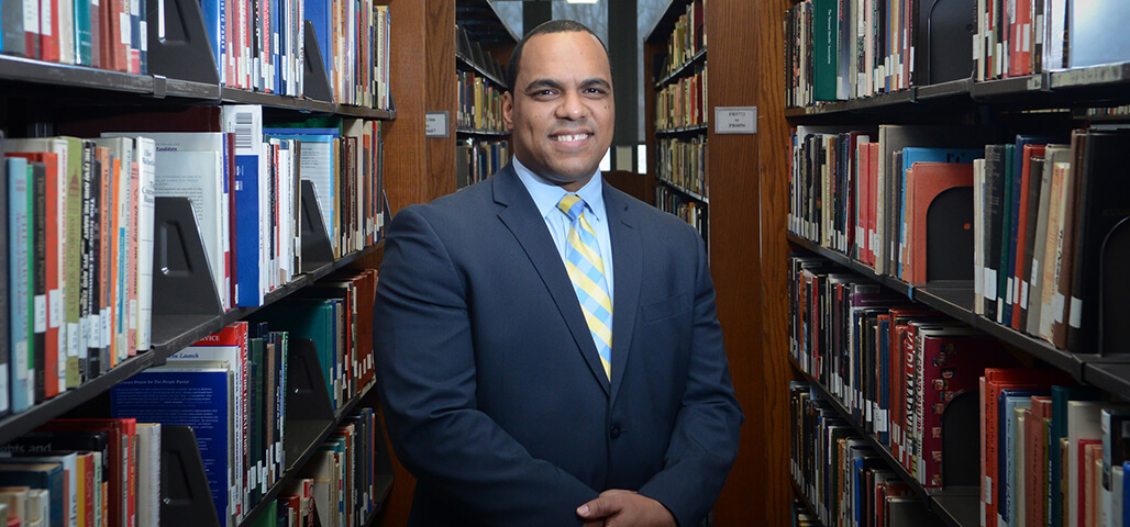 A Business Transfer Associate  Degree student dressed in formal business suit and tie, standing in the library surrounded by book shelves.