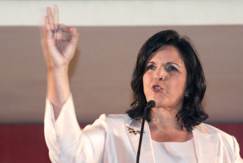 Minou speaking passionately into a microphone and raising her hand in the air.