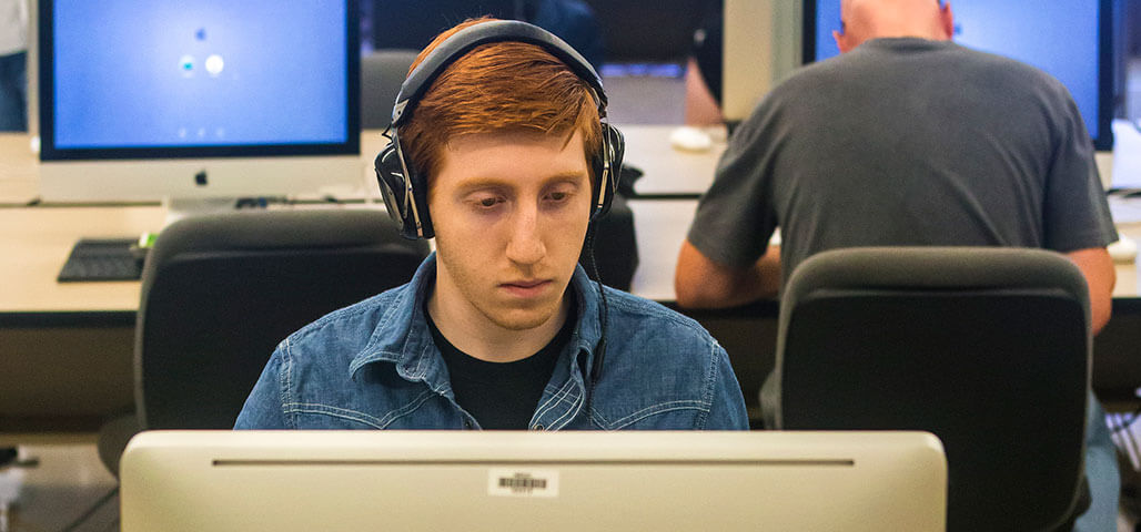 An Information Technology student sits at a computer with headphones on, engrossed in his work.