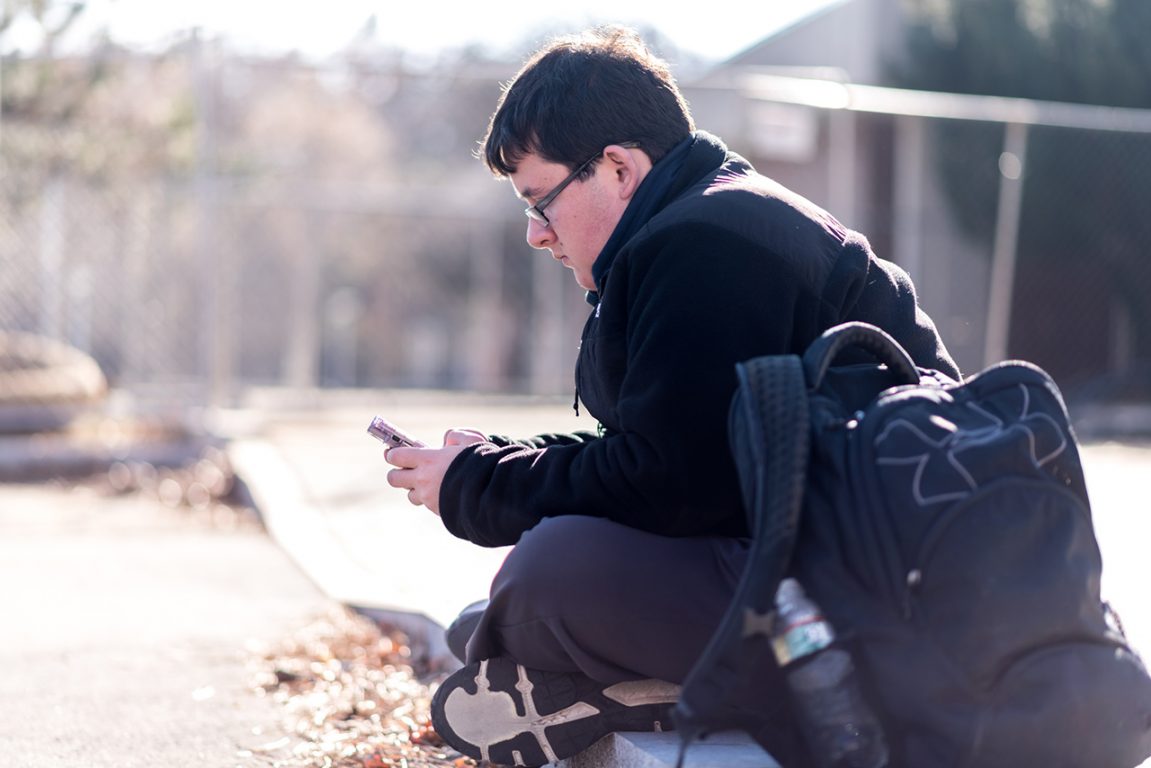 Brian Oppedisano, sitting on the ground outside, looking at his phone.