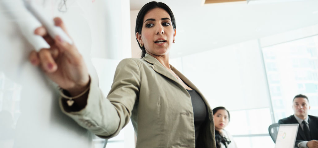 A woman at a meeting giving a demonstration at a whiteboard, while other employees look on.