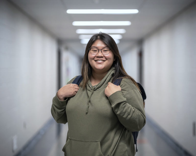 Jenni Kim, in a hallway on campus, smiling, hands supporting backpack on back.