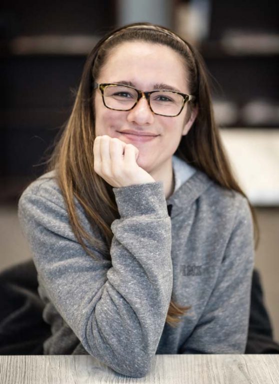 Maeina, a young woman with glasses and long hair, leans on her hand, arm propped on elbow on table.