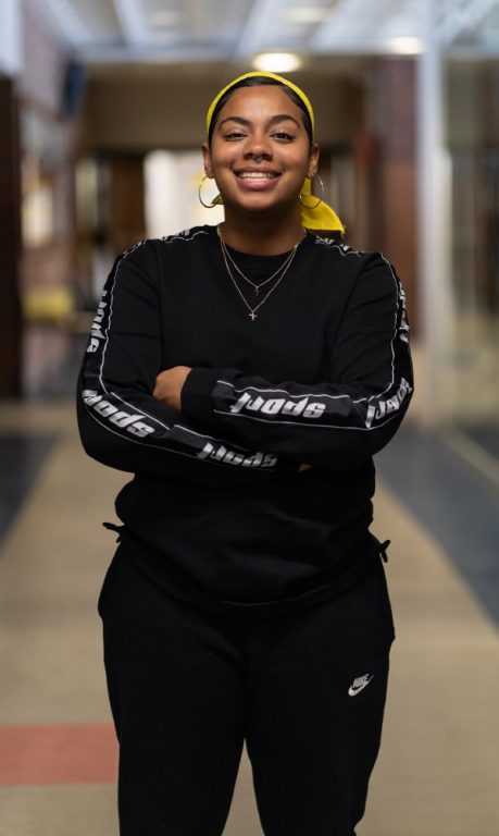 Yuliana Sanchez standing in a corridor with arms crossed over her stomache, and big smile on her face.