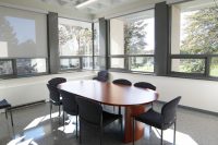 An oblong rounded table surrounded by chairs in a room with windows lining the far walls, which let in a lot of sunshine. 