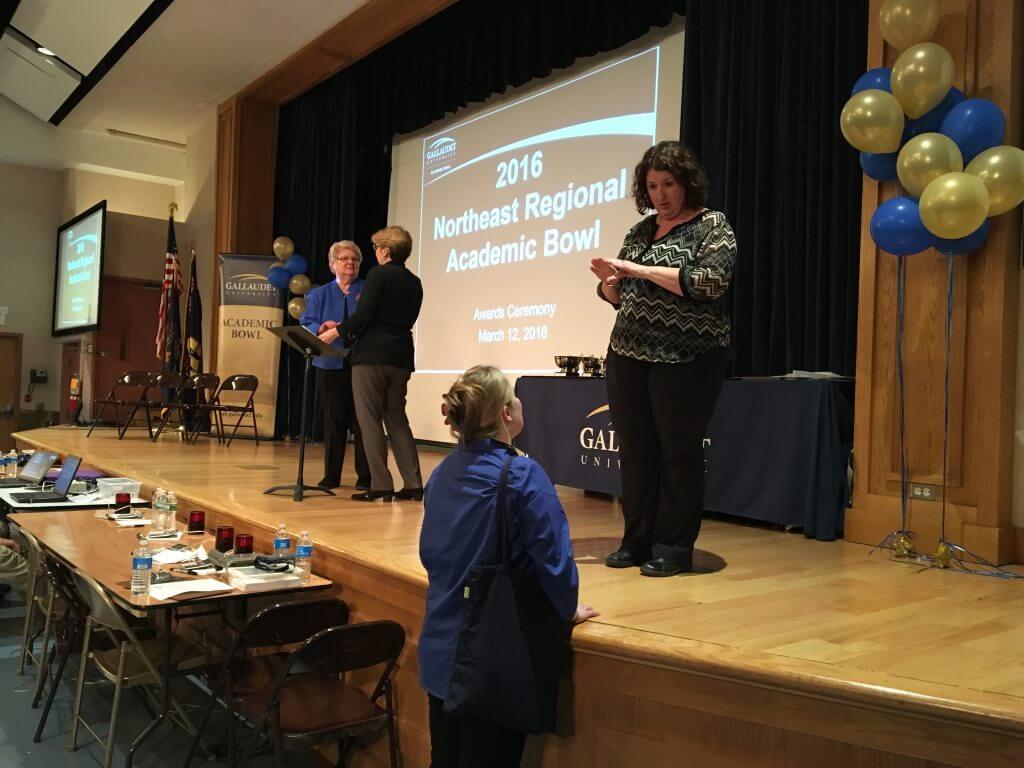 Gallaudet University Regional Center East, host school staff, and Bobbie Cordano, stand on a stage, interacting, and preparing to present the 2016 Northeast Regional Academic Bowl awards. There are balloons in Gallaudet colors, banners and a power point being projected onto two big screen