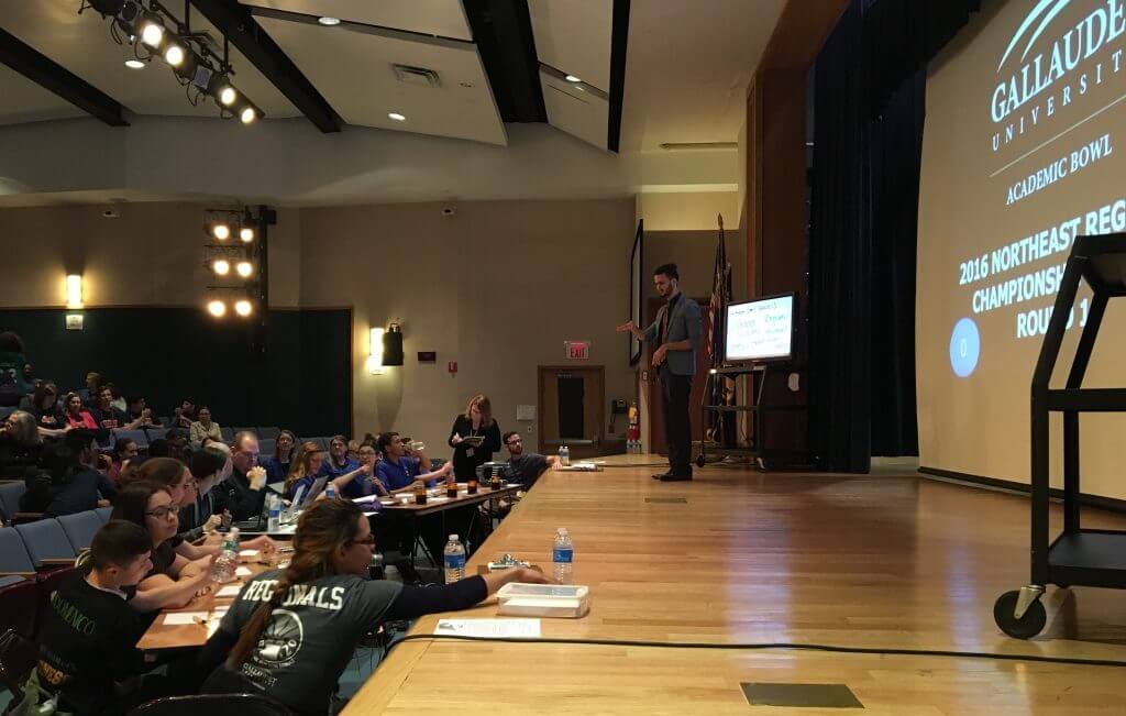 The stage at the 2016 Northeast Regional Academic Bowl Championships. A moderator is addressing the two competing teams at table in front of the stage. The students on the teams appear to be answering one of the questions. An audience watches.