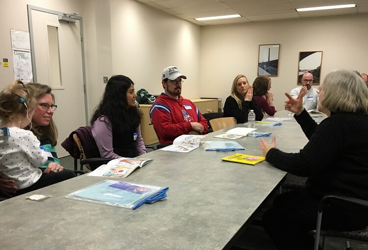 A group of adults are sitting at a table in a classroom. They all have picture books open in front of them. A tutor is in front of them signing for them.