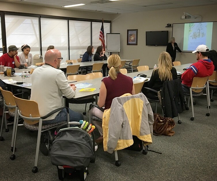 parents and guardians seated at a large horse shoe arrangement of tables watching the Deaf instructor, who is teaching them how to sign a story.