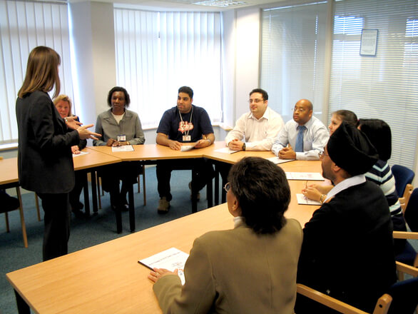 A female presenter with long hair addresses a horseshoe table full of professionals