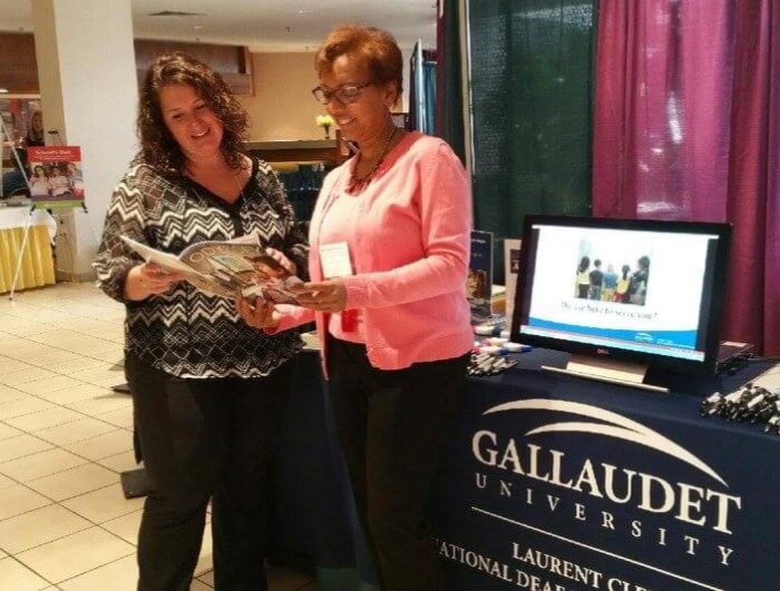GURC staff Aimee Stevens stands at a Gallaudet table at a conference. She is looking at Odessey magazine with another woman