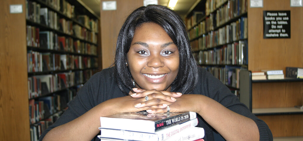 A Business Management: Healthcare Practice associate degree student in the library, smiling and leaning on a staff of books.