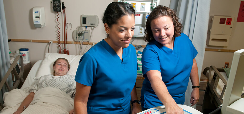 Two students in the Medical Assistant Certificate program work on equipment in a mock emergency room, while another student acts as a patient in the bed.