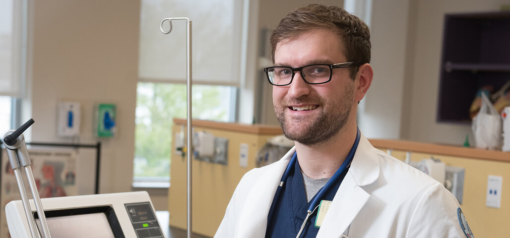 A male respiratoy care associate degree student stands by a monitor next to a bed.