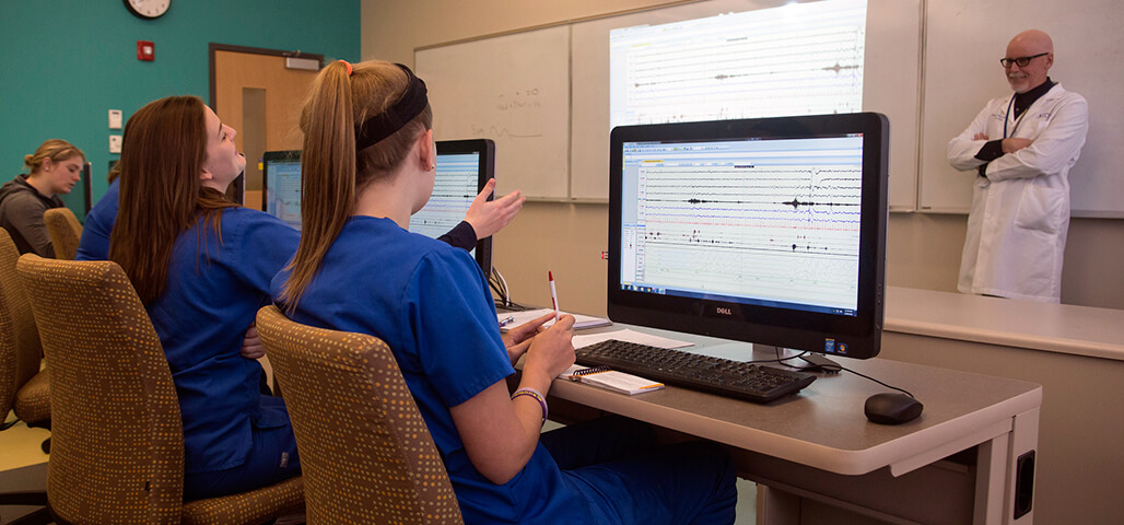 Several Sleep Technologist certificate students in a classroom looking at brain waves from a Polysomnography record on their computers, or actively engaged in conversation with the professor. 