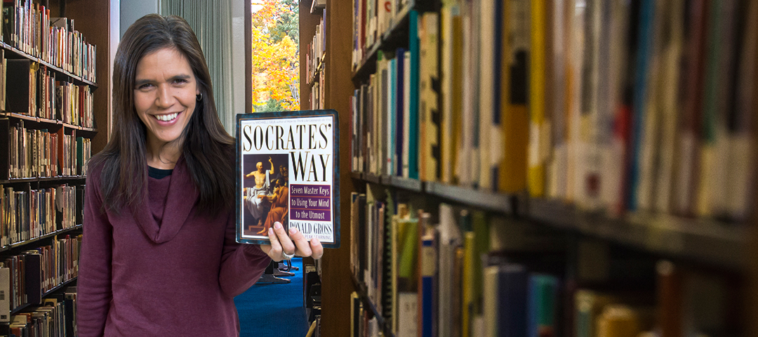 A professor that teaches courses for the Liberal Arts Philosophy degree, in the library holding up the book 'Socrates Way'.