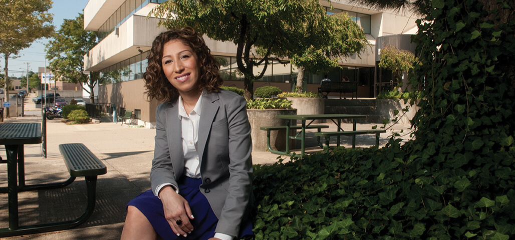 A female Liberal Arts associate degree student in formal suit and blouse, sits on a bench under a tree in front of the Dimitry building in Lawrence.