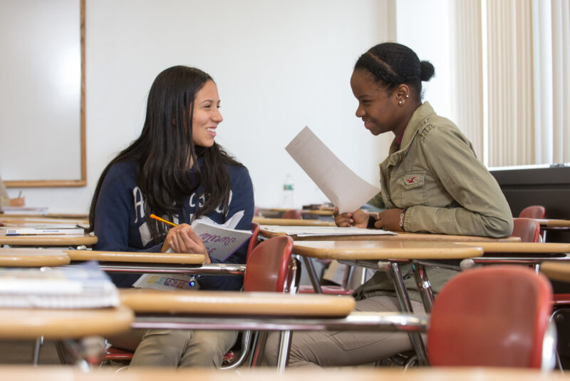 Two female presenting students chatting it up in a classroom setting
