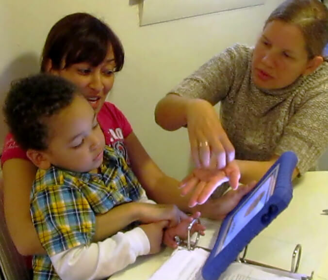 A boy, his mother, and a teacher are seated at a table. The boy is in his mother's lap. They are all looking at an iPad together. There is a picture of a cookie on the screen and the tutor is showing them the sign.